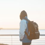 Back view of a hooded teenager with a backpack looking at the sea at sunset
