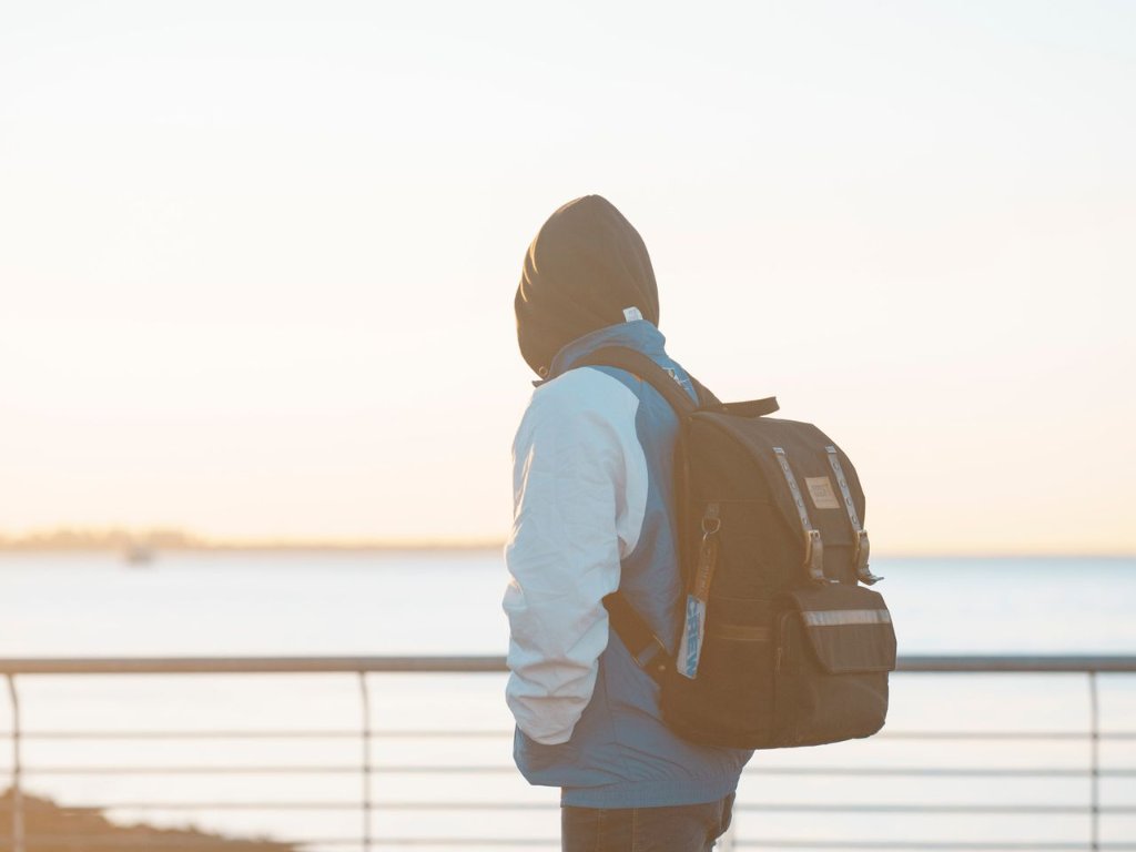 Back view of a hooded teenager with a backpack looking at the sea at sunset