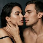 A couple at kiss distance in warm lamplight, her hand at his jaw — sexual communication and intimacy