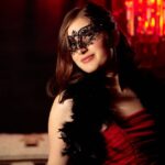 Woman in a black lace masquerade mask and red dress under red chandelier light, illustrating BDSM roles and power exchange