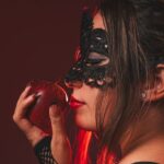 فانتزی سلطه و تسلیم؛ زن با ماسک توری مشکی و رژ قرمز سیب سرخ را نزدیک لب گرفته است
