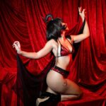 Is BDSM a Sign of Childhood Trauma? What Research Shows زن جوان با لباس زیر قرمز و ماسک نقابدار روی پردهٔ مخمل قرمز، نماد نقشپذیری در BDSM و پرسش رابطهٔ آن با ترومای کودکی